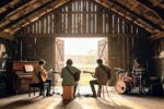 Quatre musiciens jouant de la guitare et de la batterie dans une grange rustique face à un champ ensoleillé